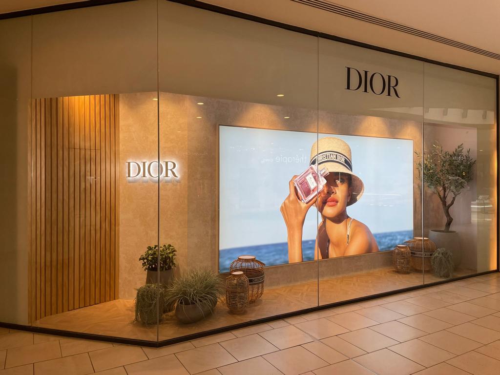 Dior store front with a display of a woman holding a product, surrounded by plants.