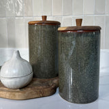 Two dark turquoise ceramic canisters with wooden lids on a white surface with tiled wall background. Interior decor. Rtfact