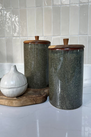 Two dark turquoise ceramic canisters with wooden lids on a white surface with tiled wall background. Interior decor. Rtfact