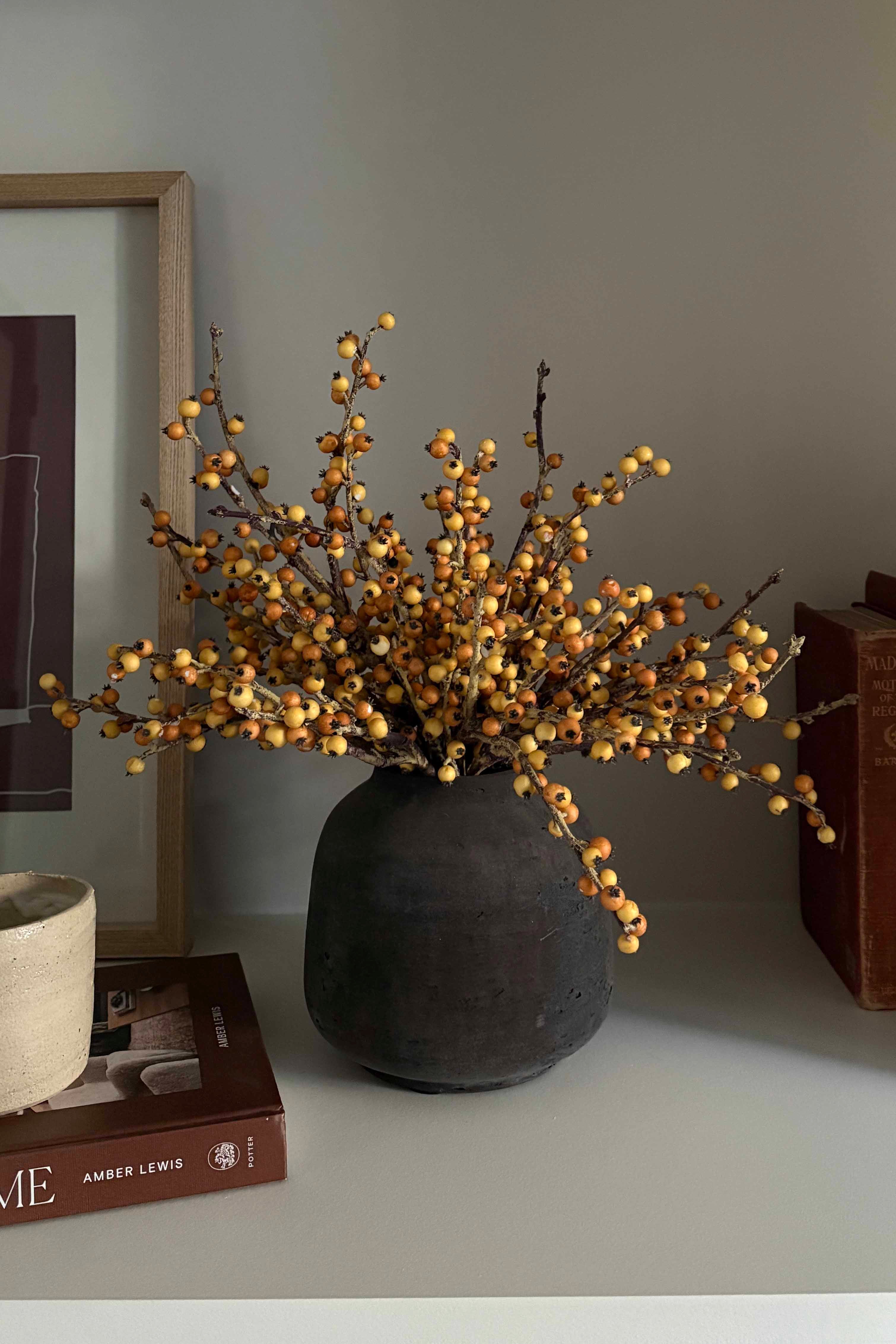 Decorative faux plant in a black vase on a shelf with books and a framed picture. Rtfact Flowers
