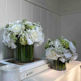 Two faux floral arrangements in glass vases on a white surface with a neutral background. Rtfact