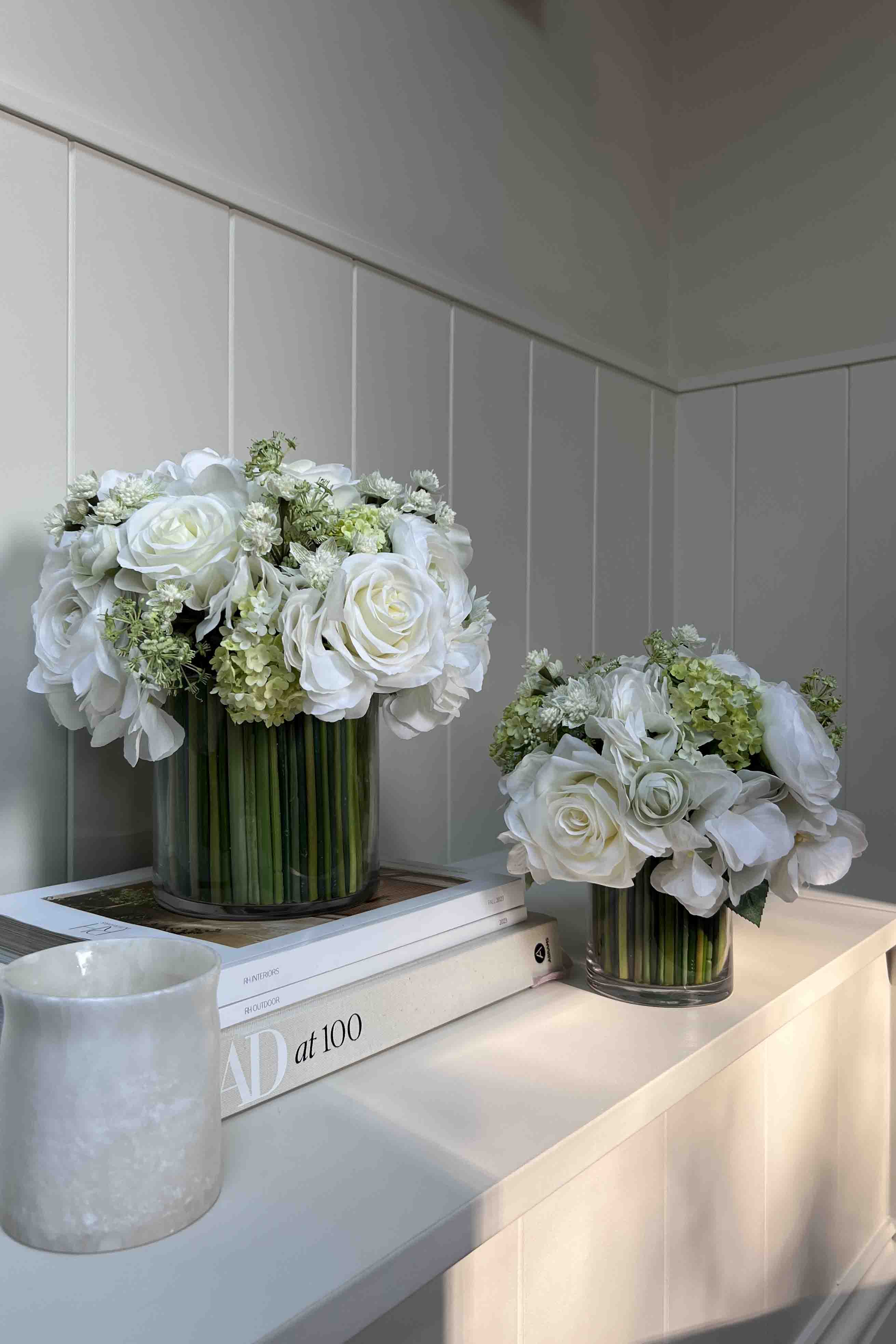 Two faux floral arrangements in glass vases on a white surface with a neutral background. Rtfact