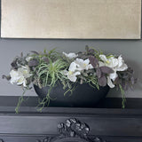 A faux floral arrangement featuring white orchids and green plants in a black stone-like boat, placed on a dark surface with a wall in the background.