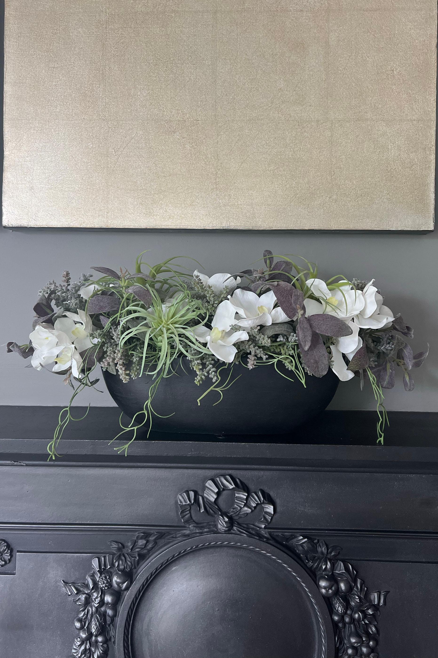 A faux floral arrangement featuring white orchids and green plants in a black stone-like boat, placed on a dark surface with a wall in the background.