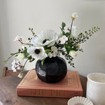 Faux white Floral arrangement in a black vase on a wooden surface with a book and small dish. Interior decor. rtfact