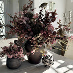 A large arrangement of burgundy Eucalyptus, Angelica, and Wax Flowers in a rustic brown stone vase, displayed in a bright room setting.