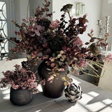 A large arrangement of burgundy Eucalyptus, Angelica, and Wax Flowers in a rustic brown stone vase, displayed in a bright room setting.