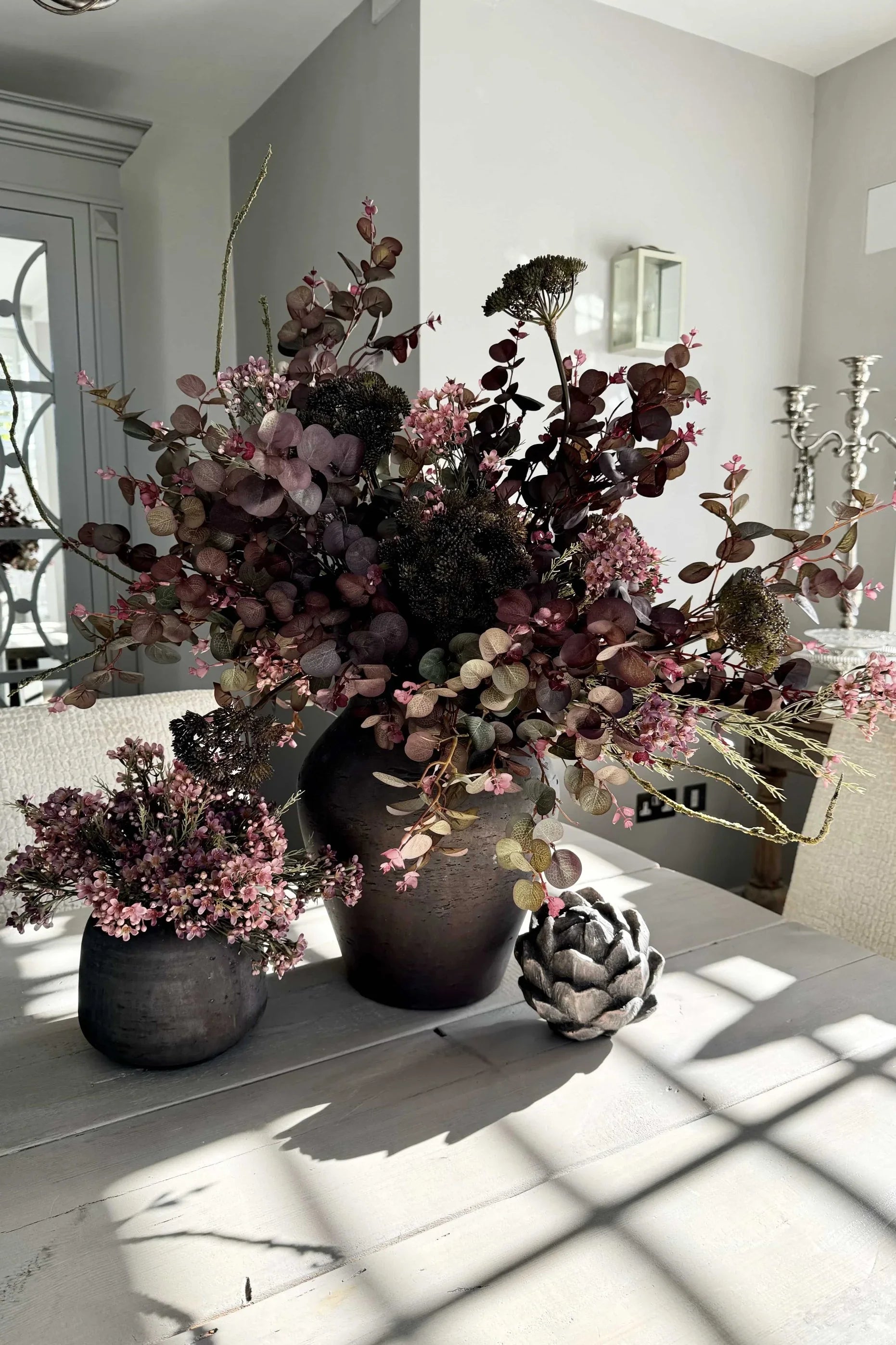 A large arrangement of burgundy Eucalyptus, Angelica, and Wax Flowers in a rustic brown stone vase, displayed in a bright room setting.
