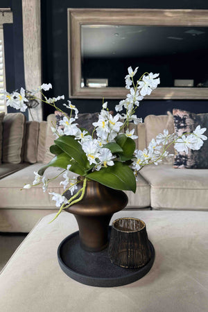 Faux arrangement of white flowers in a bronze vase on a coffee table in a living room. Rtfact
