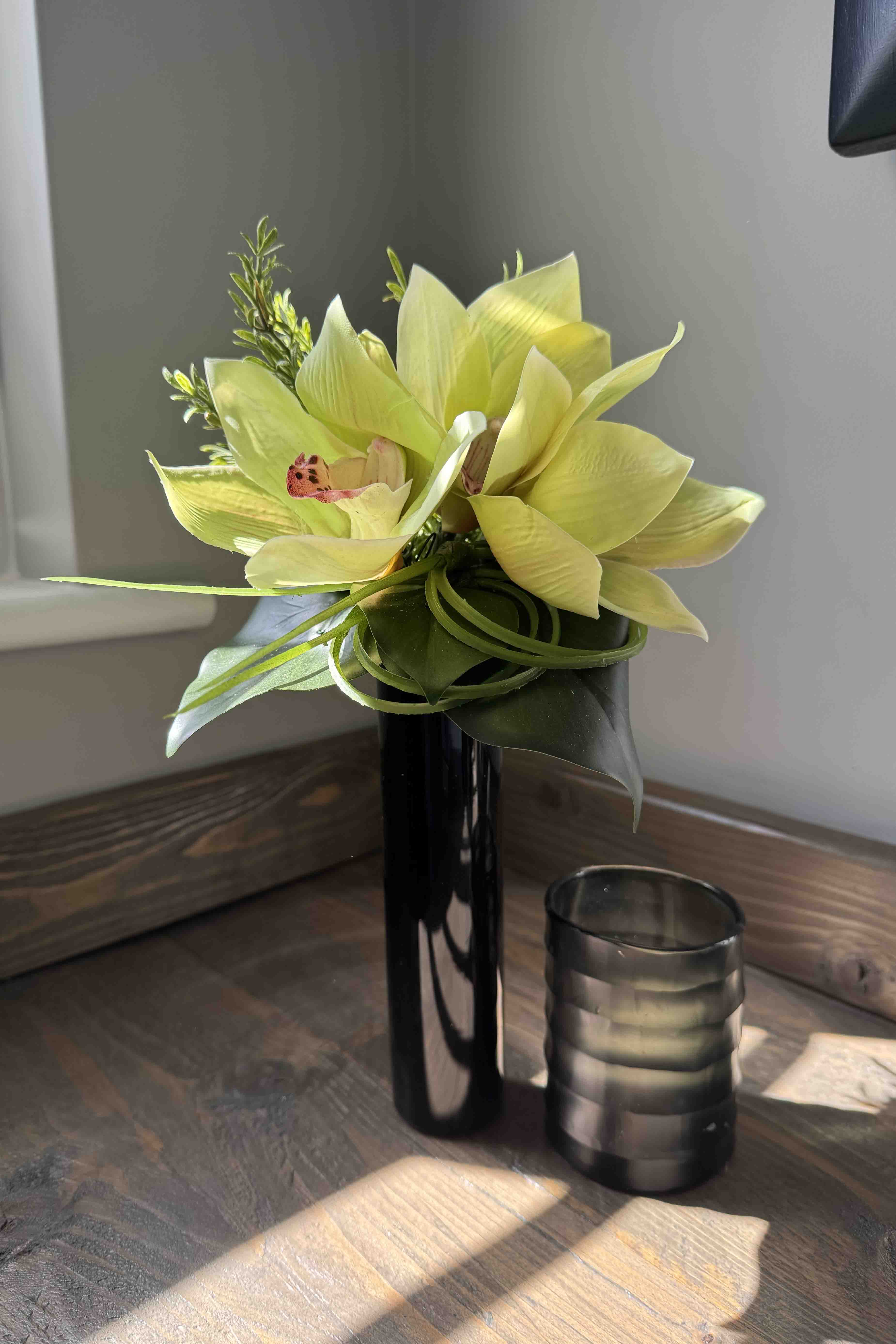 Vase with faux orchid flowers on a wooden surface next to a glass container. Rtfact 