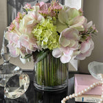 Faux bouquet of flowers in a clear vase on a reflective surface with decorative items in the background.