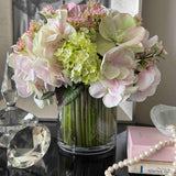 Faux bouquet of flowers in a clear vase on a reflective surface with decorative items in the background.