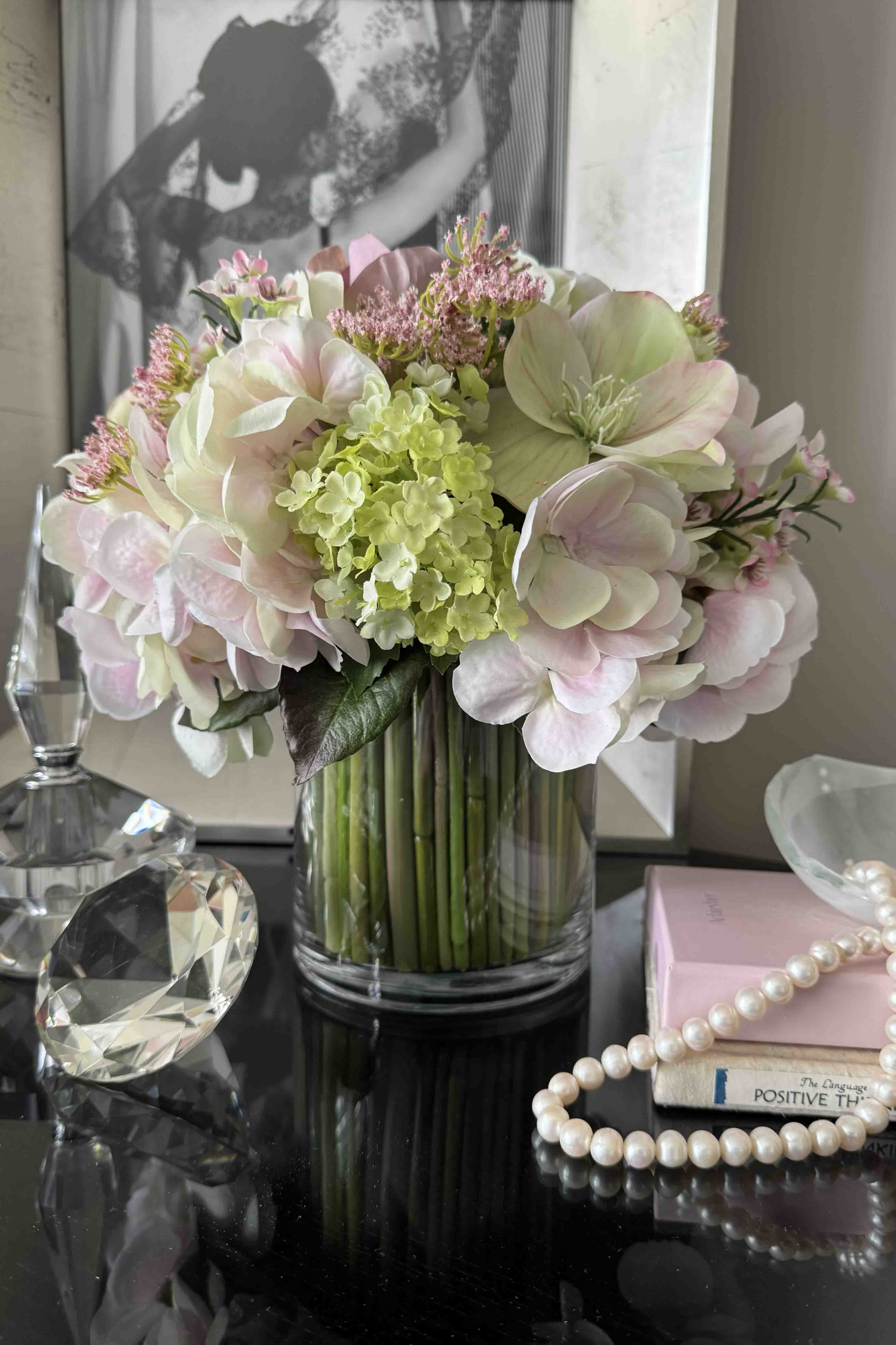 Faux bouquet of flowers in a clear vase on a reflective surface with decorative items in the background.