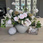 A floral arrangement of soft pink roses, hydrangea, and astrantia in a rustic light stone color vase, accompanied by greenery.