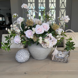 A floral arrangement of soft pink roses, hydrangea, and astrantia in a rustic light stone color vase, accompanied by greenery.
