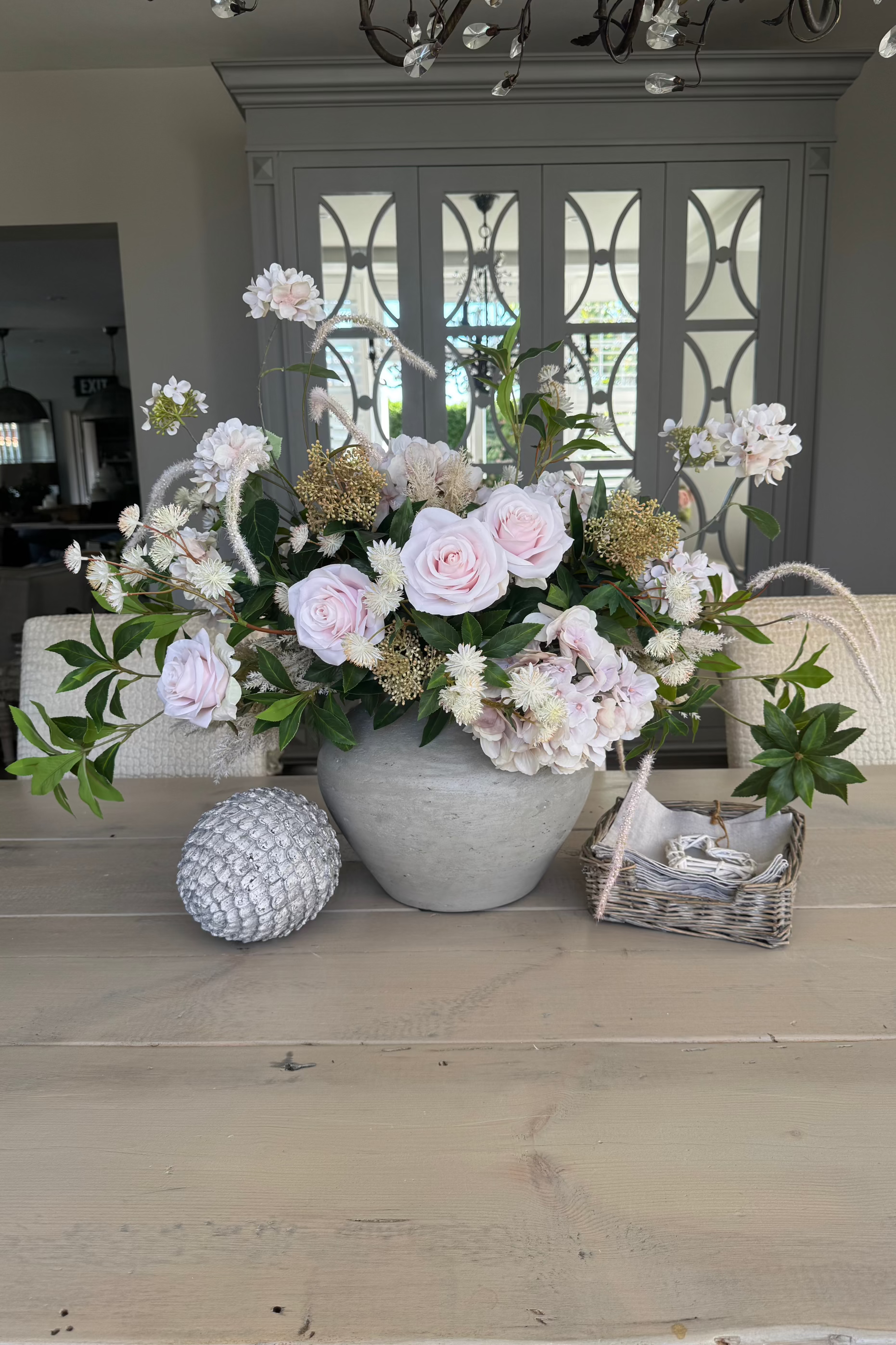 A floral arrangement of soft pink roses, hydrangea, and astrantia in a rustic light stone color vase, accompanied by greenery.