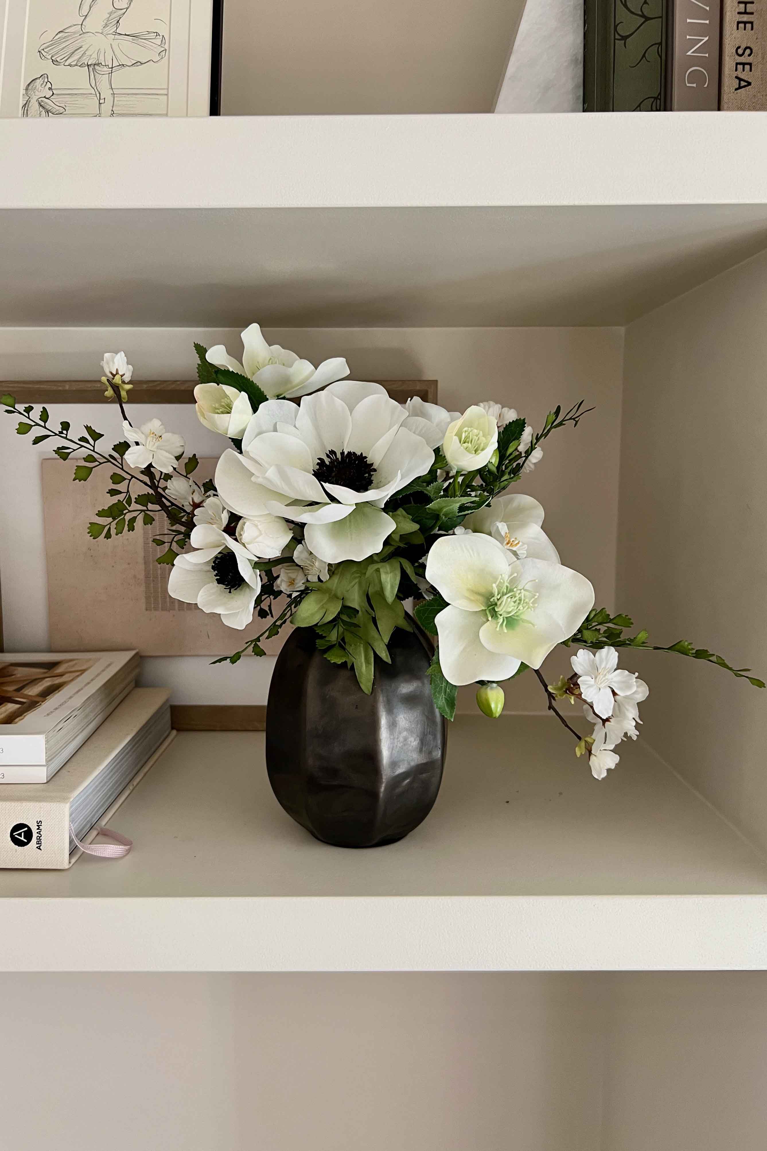Faux Floral arrangement in a black vase on a shelf with books and decorative items.