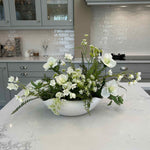 Faux floral arrangement in a white vase on a kitchen counter with cabinets and appliances in the background.
