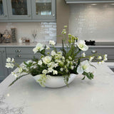 Faux floral arrangement in a white vase on a kitchen counter with cabinets and appliances in the background.