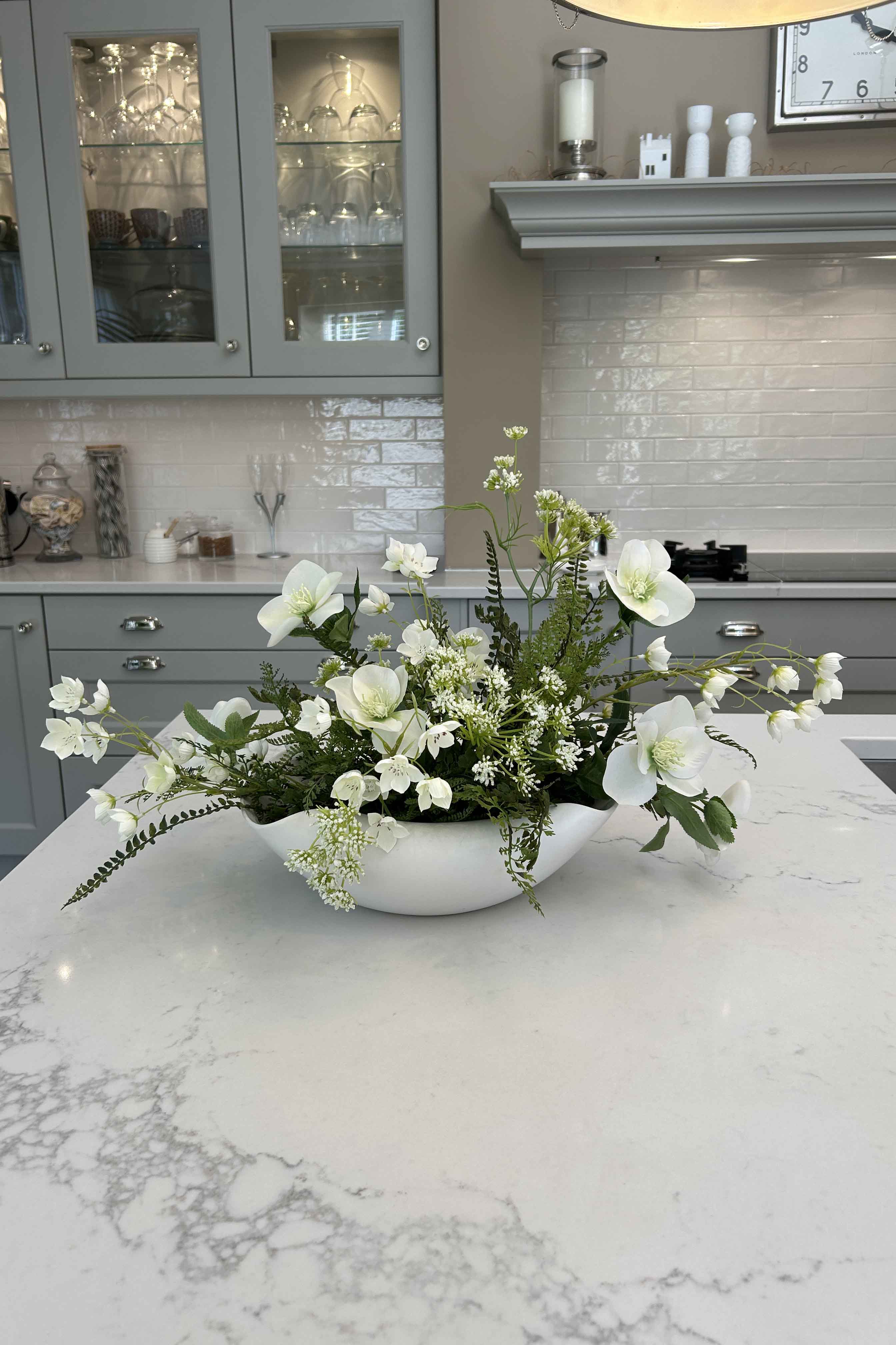 Faux floral arrangement in a white vase on a kitchen counter with cabinets and appliances in the background.