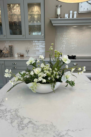 Faux floral arrangement in a white vase on a kitchen counter with cabinets and appliances in the background.
