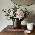 Faux floral arrangement in a vase on a wooden surface with a book and candle.