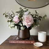 Faux floral arrangement in a vase on a wooden surface with a book and candle.