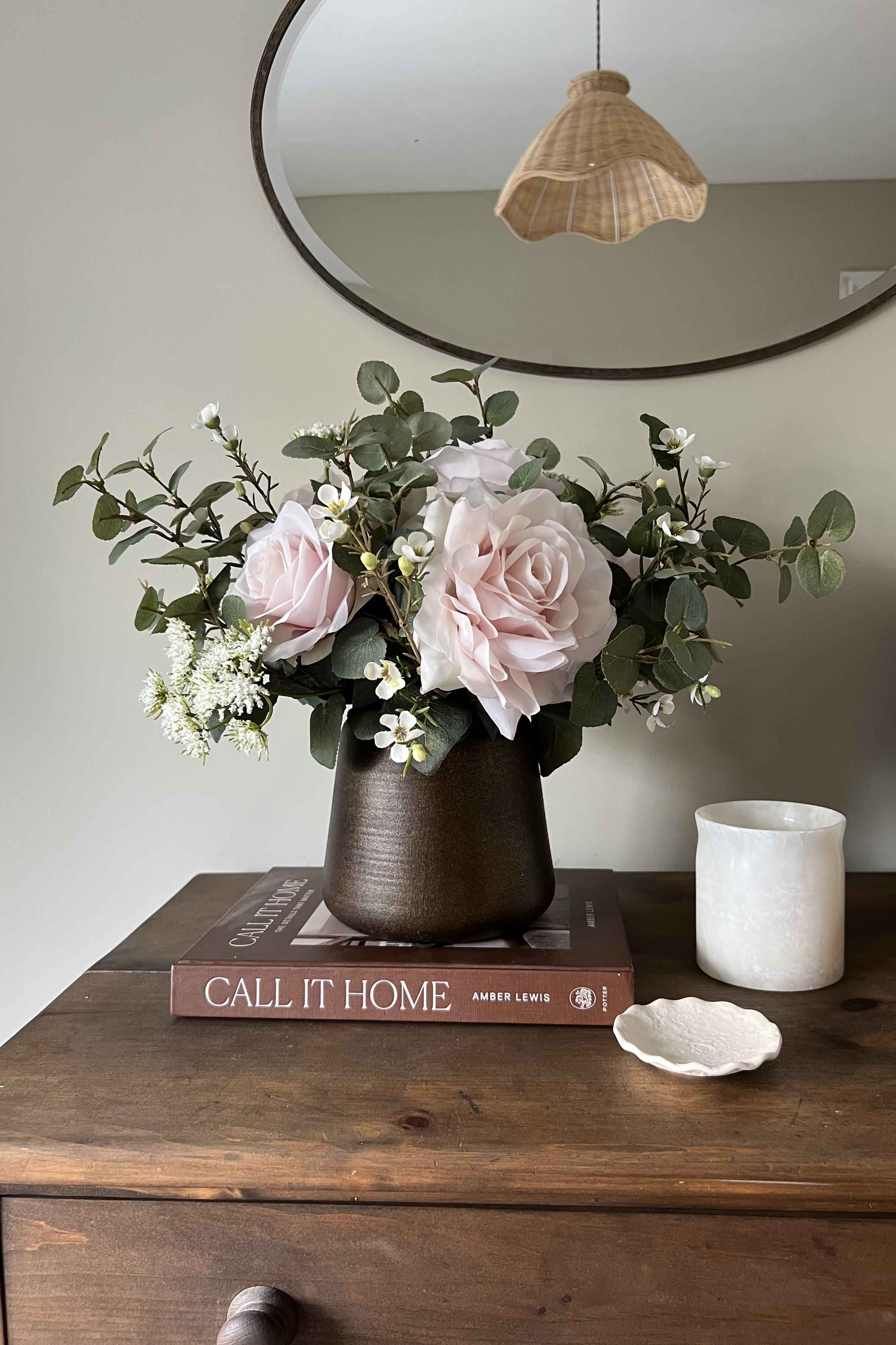 Faux floral arrangement in a vase on a wooden surface with a book and candle.
