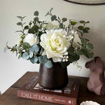Faux floral arrangement in a dark vase on a wooden surface with books and a decorative object.