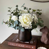 Faux floral arrangement in a dark vase on a wooden surface with books and a decorative object.