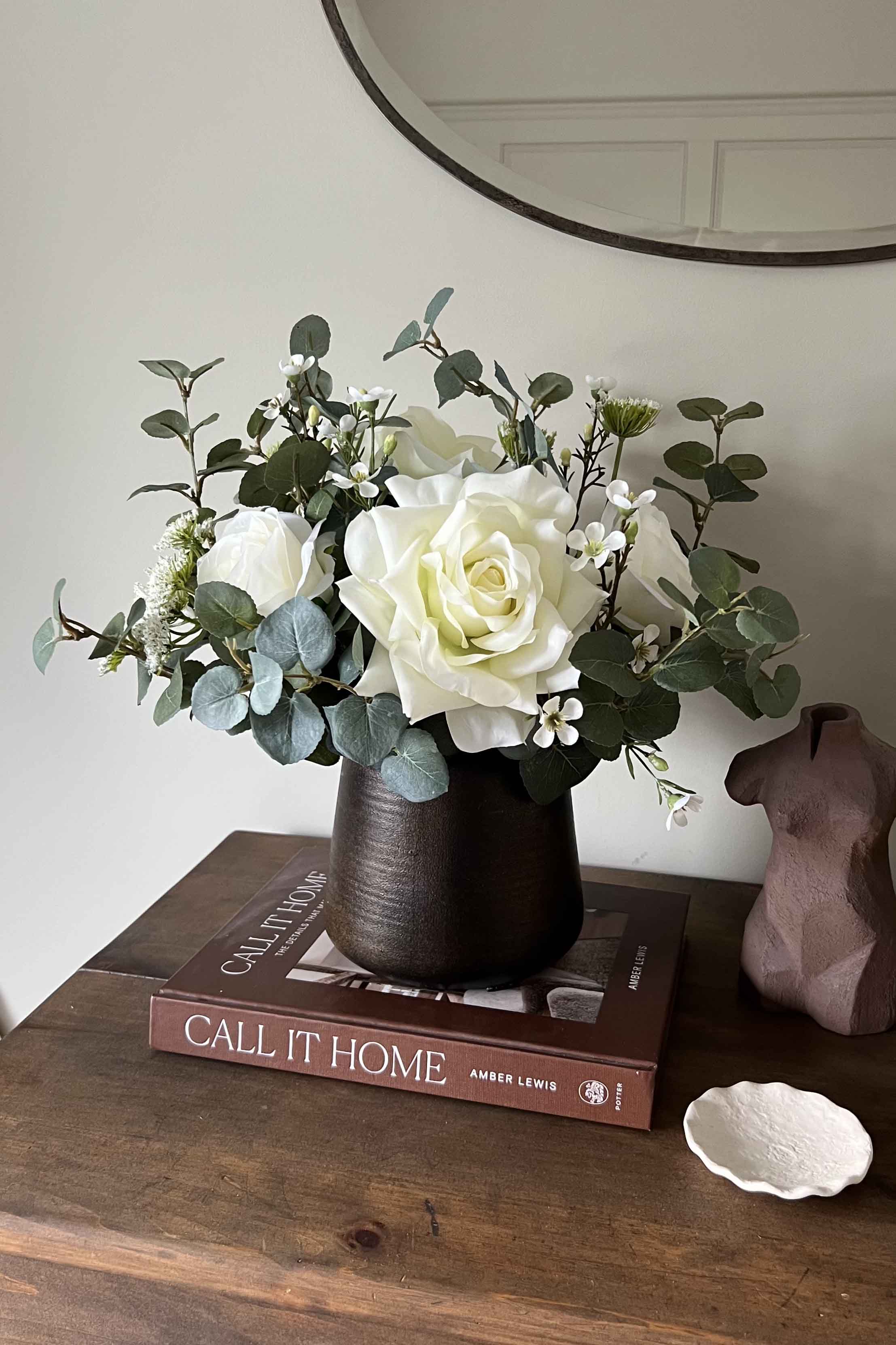 Faux floral arrangement in a dark vase on a wooden surface with books and a decorative object.