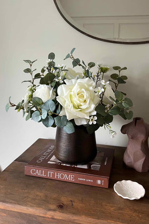 Faux floral arrangement in a dark vase on a wooden surface with books and a decorative object.