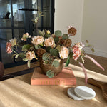Decorative faux floral arrangement on a wooden surface with a blurred background. rtfact