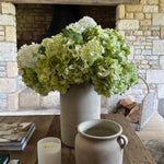 Decorative arrangement with faux flowers, candle, and pot on a wooden table in front of a stone fireplace. rtfact
