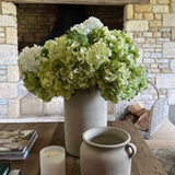 Decorative arrangement with faux flowers, candle, and pot on a wooden table in front of a stone fireplace. rtfact