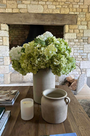 Decorative arrangement with faux flowers, candle, and pot on a wooden table in front of a stone fireplace. rtfact