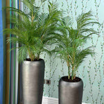 Two tall gray planters with faux green plants against a light blue wall.