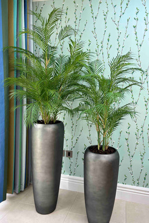 Two tall gray planters with faux green plants against a light blue wall.