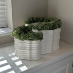 Three white textured planters with faux green plants on a white surface. Rtfact