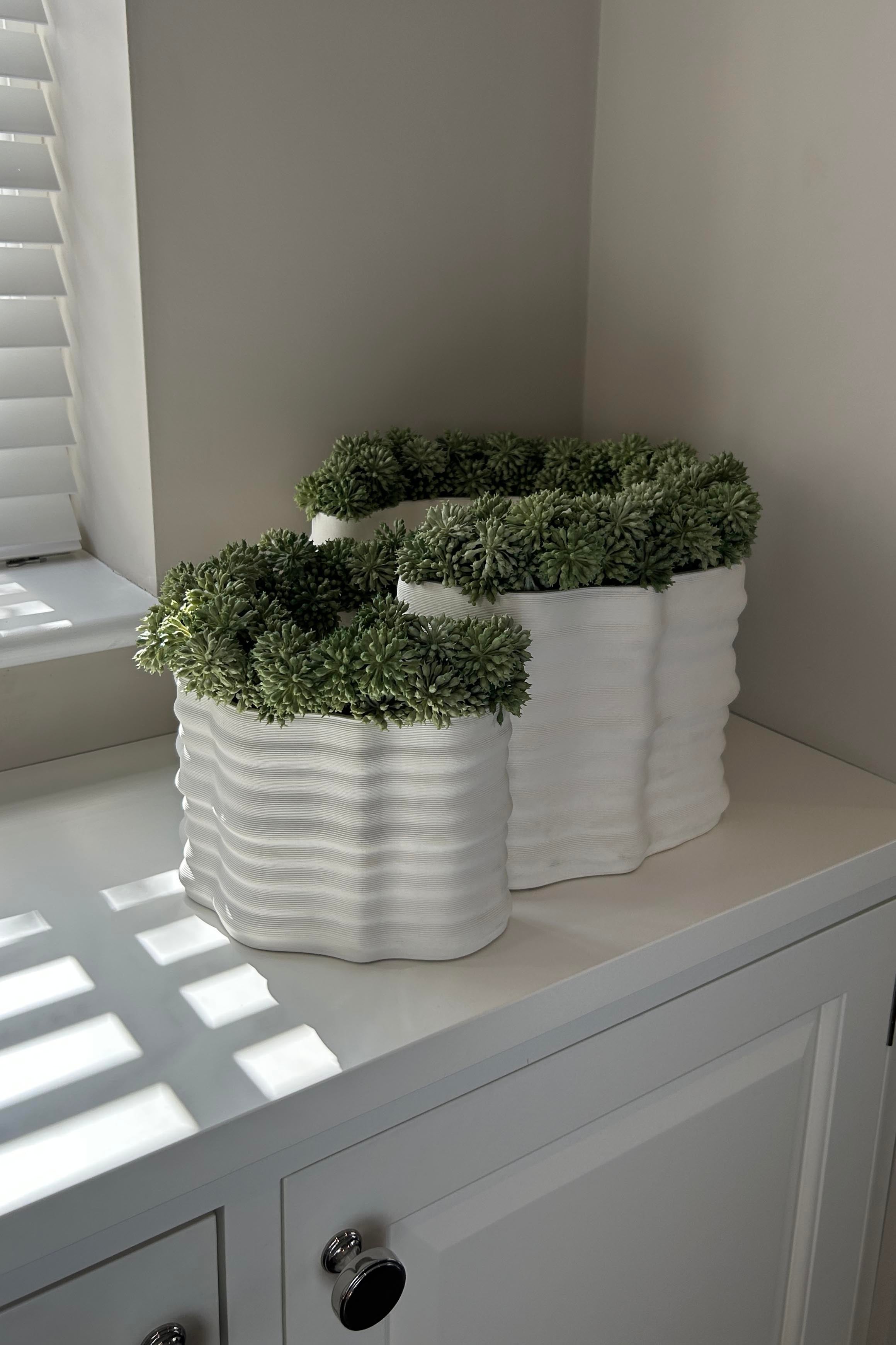 Three white textured planters with faux green plants on a white surface. Rtfact