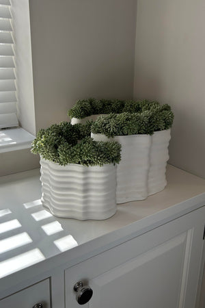 Three white textured planters with faux green plants on a white surface. Rtfact