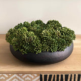Green decorative ball in a black bowl on a wooden surface with a patterned wall in the background