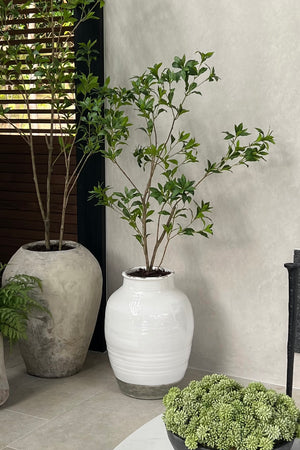 Artificial tree in a white vase with green plant against a neutral wall