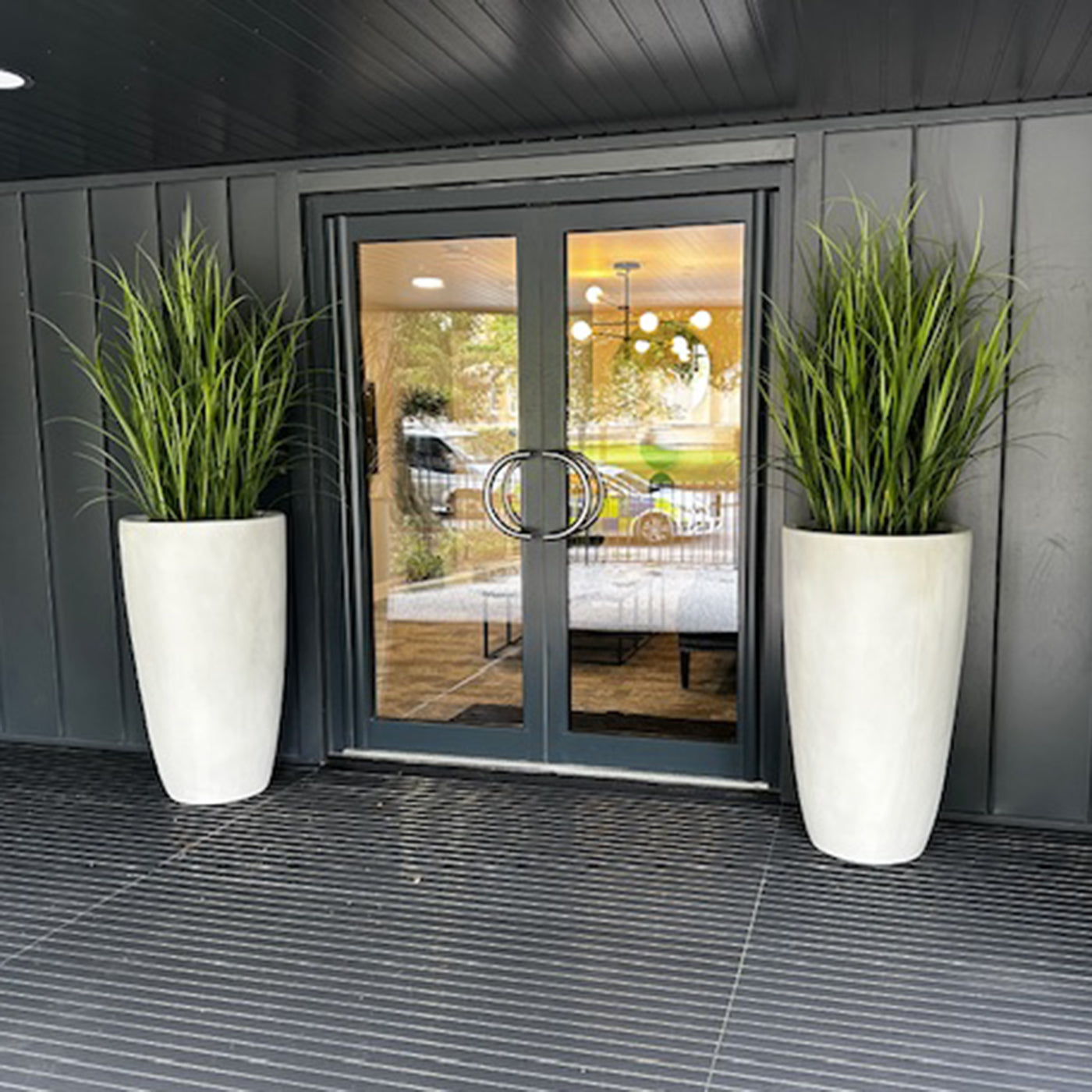 Two tall white planters with green plants on a patio area with a glass door in the background.